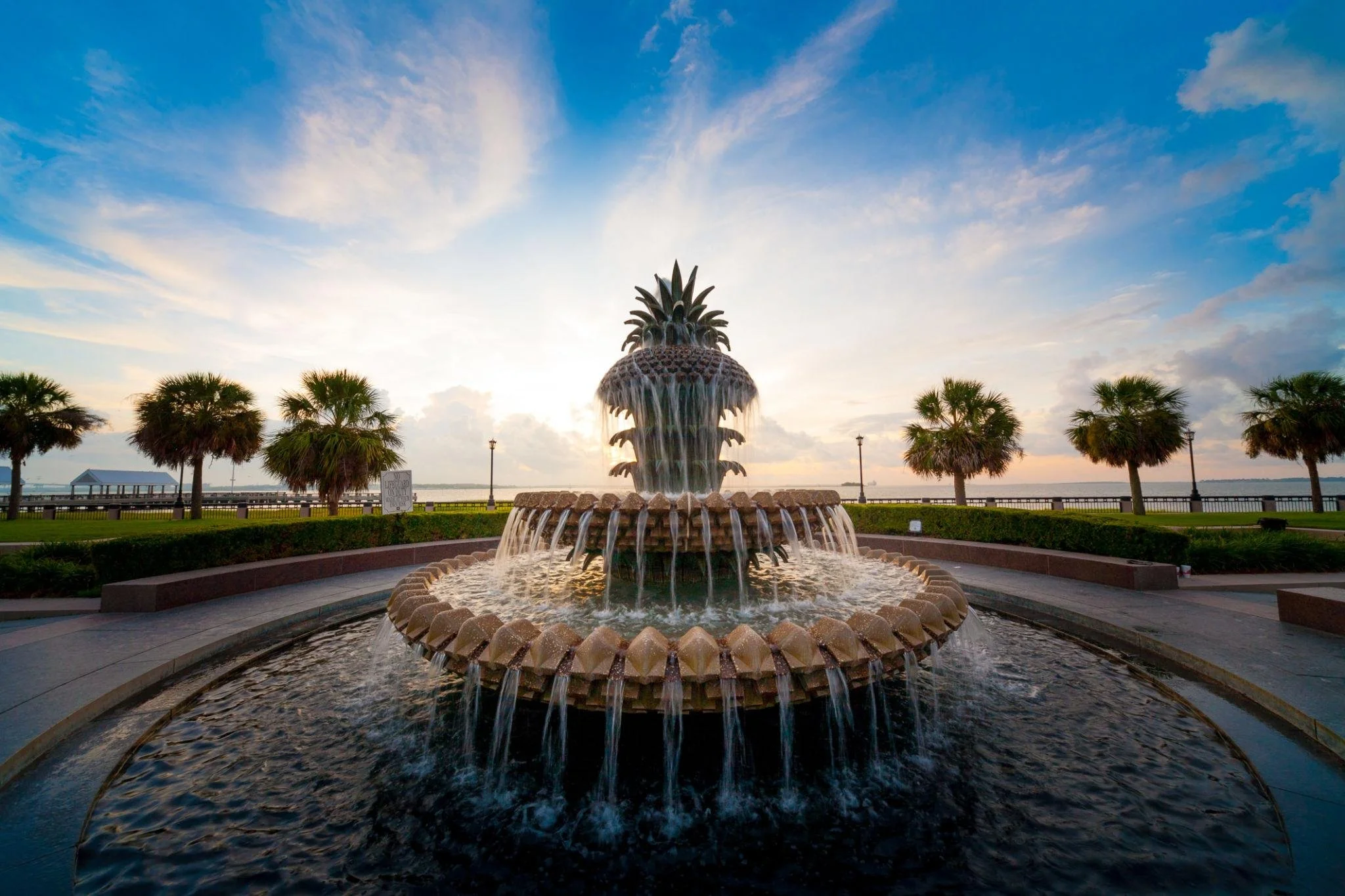 A large decorative fountain shaped like a pineapple with water flowing down its sides, set in a landscaped park with palm trees, a gazebo, and a view of the ocean at sunset or sunrise.