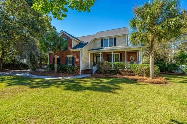 A large two-story house with a front porch, surrounded by a well-maintained lawn, trees, and palm trees under a clear blue sky.