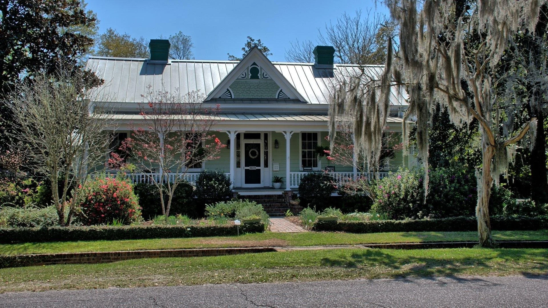 A charming green house with a metal roof, front porch, surrounded by trees and shrubs, with Spanish moss hanging from a large tree on the right.