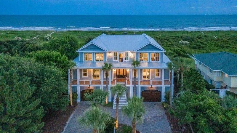 A large, multi-story beach house with a light blue roof, surrounded by palm trees, overlooking the ocean. The house is lit from inside, with a staircase leading up to the entrance.