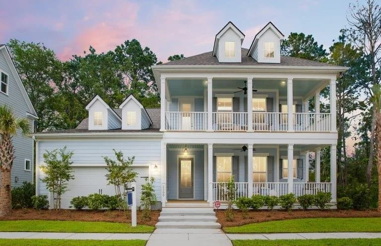 Front view of a two-story house with white siding, a covered front porch, and a two-car garage. The house has a gabled roof with three dormer windows. There are trees and shrubs around the house, and the sky is pink with clouds at sunset.
