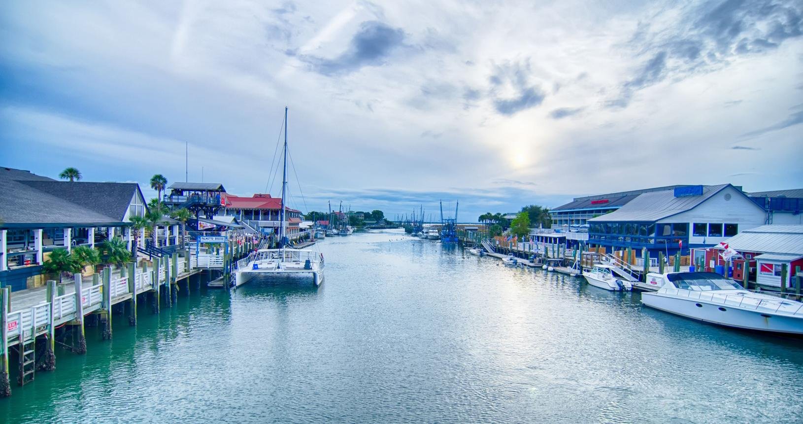 Scenic view of a marina with boats docked along wooden piers, colorful waterfront buildings, palm trees, and a partly cloudy sky.