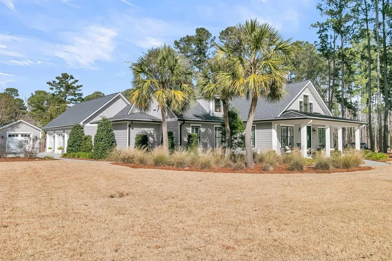 A large gray house with a covered porch, surrounded by palm trees, bushes, and a landscaped yard with yellowish-brown grass.