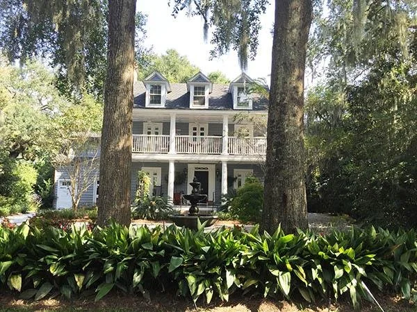 A large white two-story house with a porch and balcony, surrounded by tall trees and greenery, with a small fountain in front.