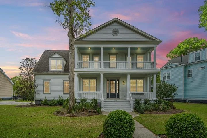 Two-story house with a front porch, white siding, black front door, lit windows, surrounded by green lawns and bushes, during sunset.