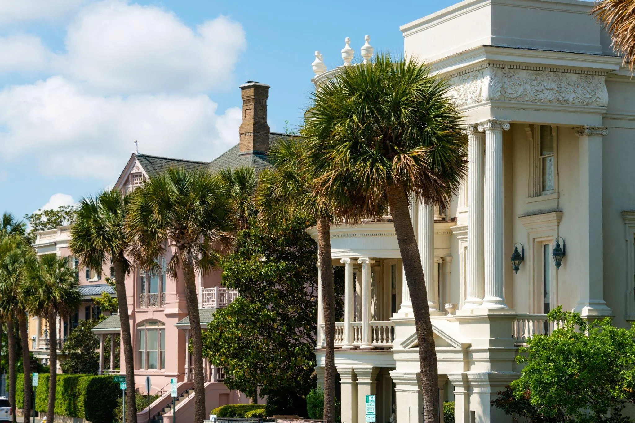 Street view of ornate white Victorian-style house with decorative trim, surrounded by tall palm trees, on a sunny day with partly cloudy sky.