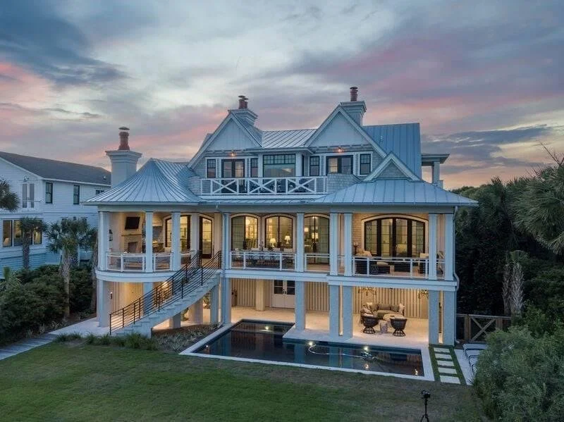 A large, multi-story house with a blue roof and white exterior, featuring an expansive porch and a swimming pool in the backyard at sunset.