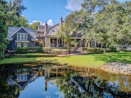 Large house with blue and gray exterior, surrounded by trees, next to a pond reflecting the house and trees.