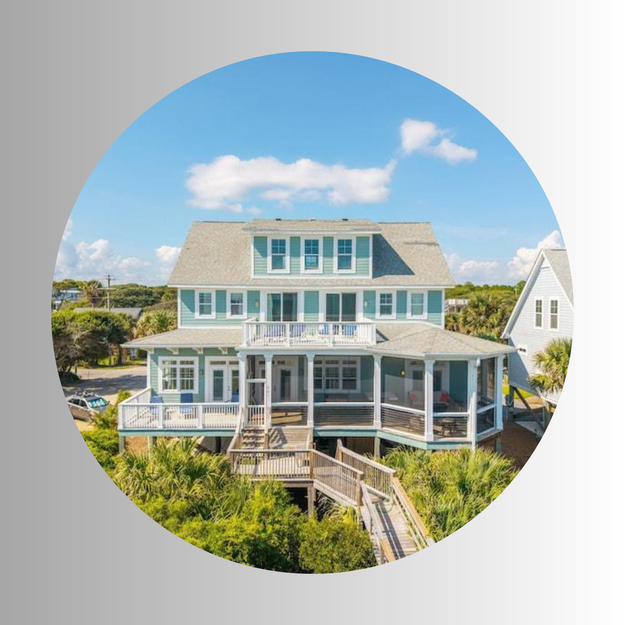 A large house with a steep gray roof, light blue siding, multiple windows, and a wraparound porch, set against a bright blue sky with scattered white clouds.