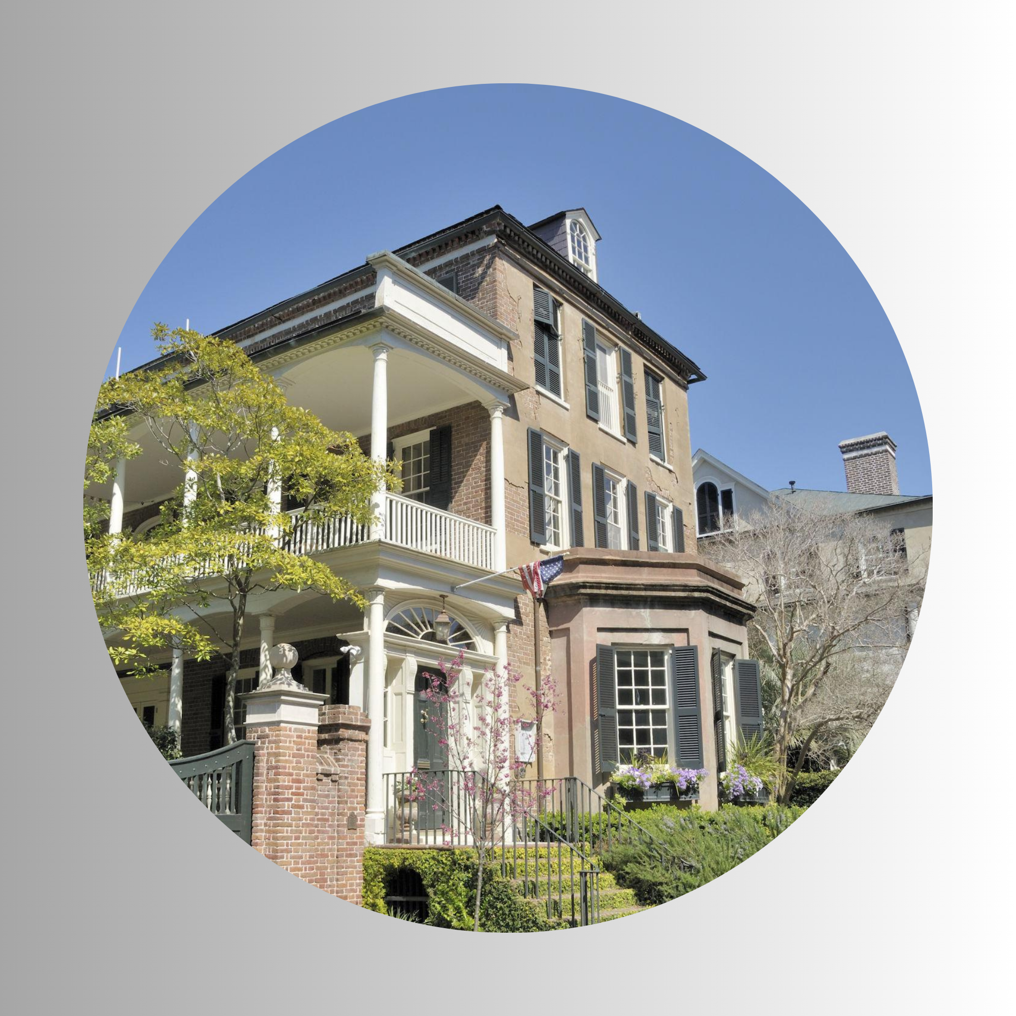 A multi-story house with brick and beige exterior, black shutters, a large front porch with white columns, surrounded by trees and greenery, under a clear blue sky.