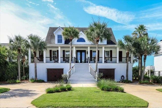 A large, white beach house with a front staircase leading to a porch, surrounded by palm trees and a manicured lawn, under a blue sky.