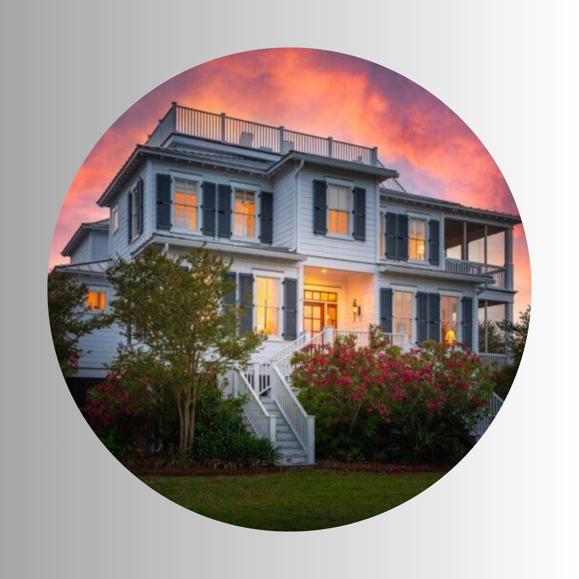 A white house with black shutters and a wrap-around porch, situated in a garden with pink flowers; sunset sky with orange and purple clouds in the background.