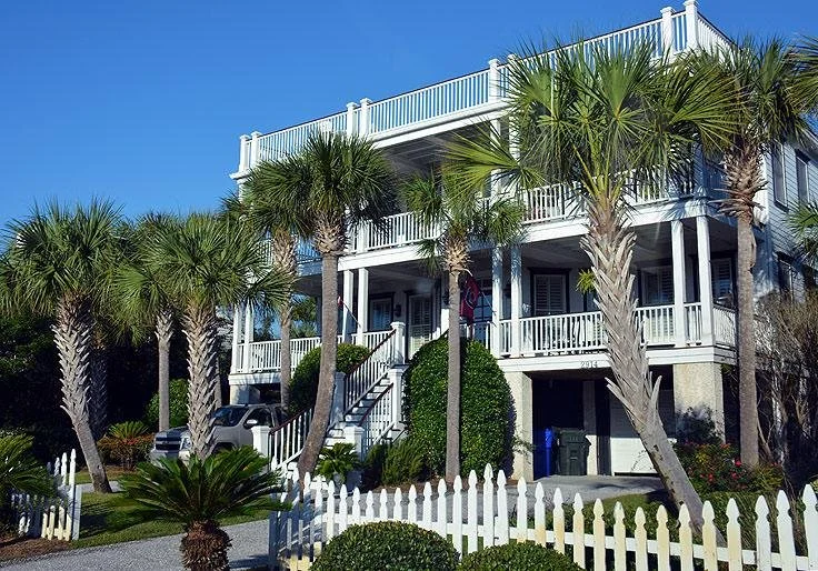 A two-story beach house with a white exterior, surrounded by palm trees and greenery, with a white picket fence in front and a clear blue sky overhead.