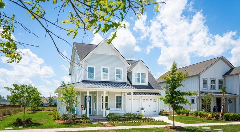A modern two-story house with white siding, a dark roof, and a front porch. The house features multiple windows, a garage, and is surrounded by a well-maintained lawn and small trees, with a bright blue sky and some clouds overhead.