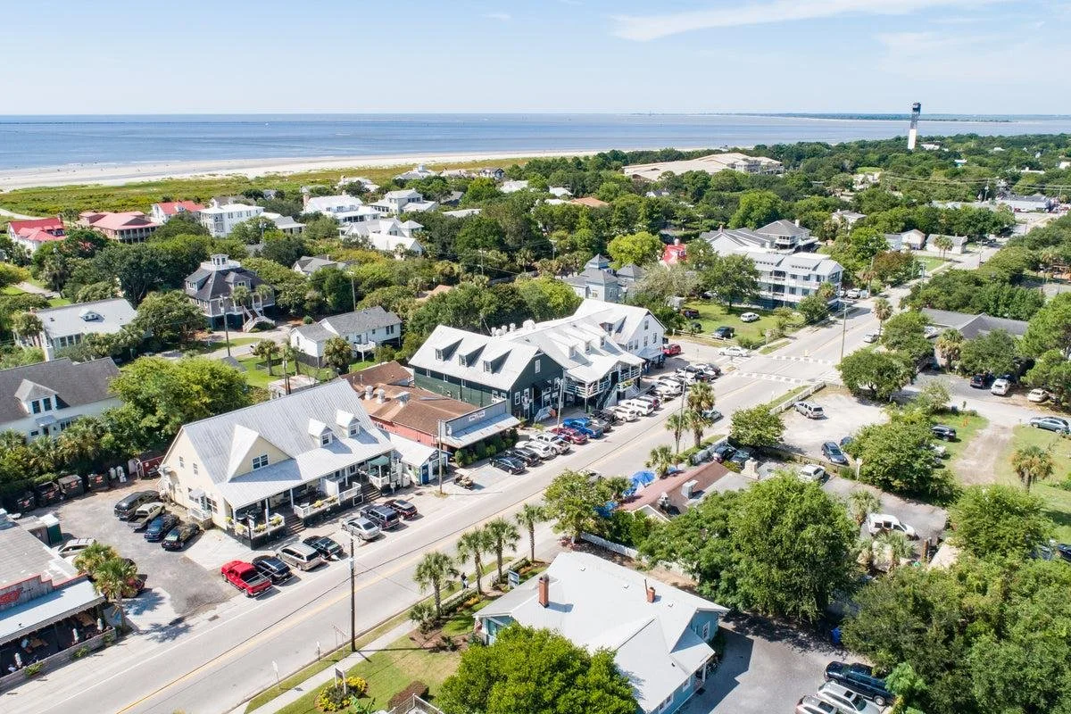 Aerial view of a coastal town with houses, trees, a street with parked cars, and the ocean in the background.