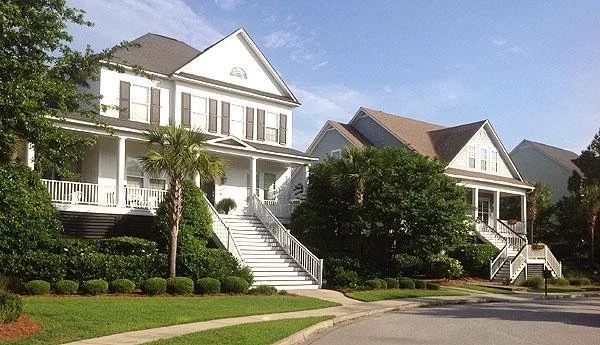Two large white houses with front porches and stairs, surrounded by manicured lawns, trees, and shrubs under a clear blue sky.