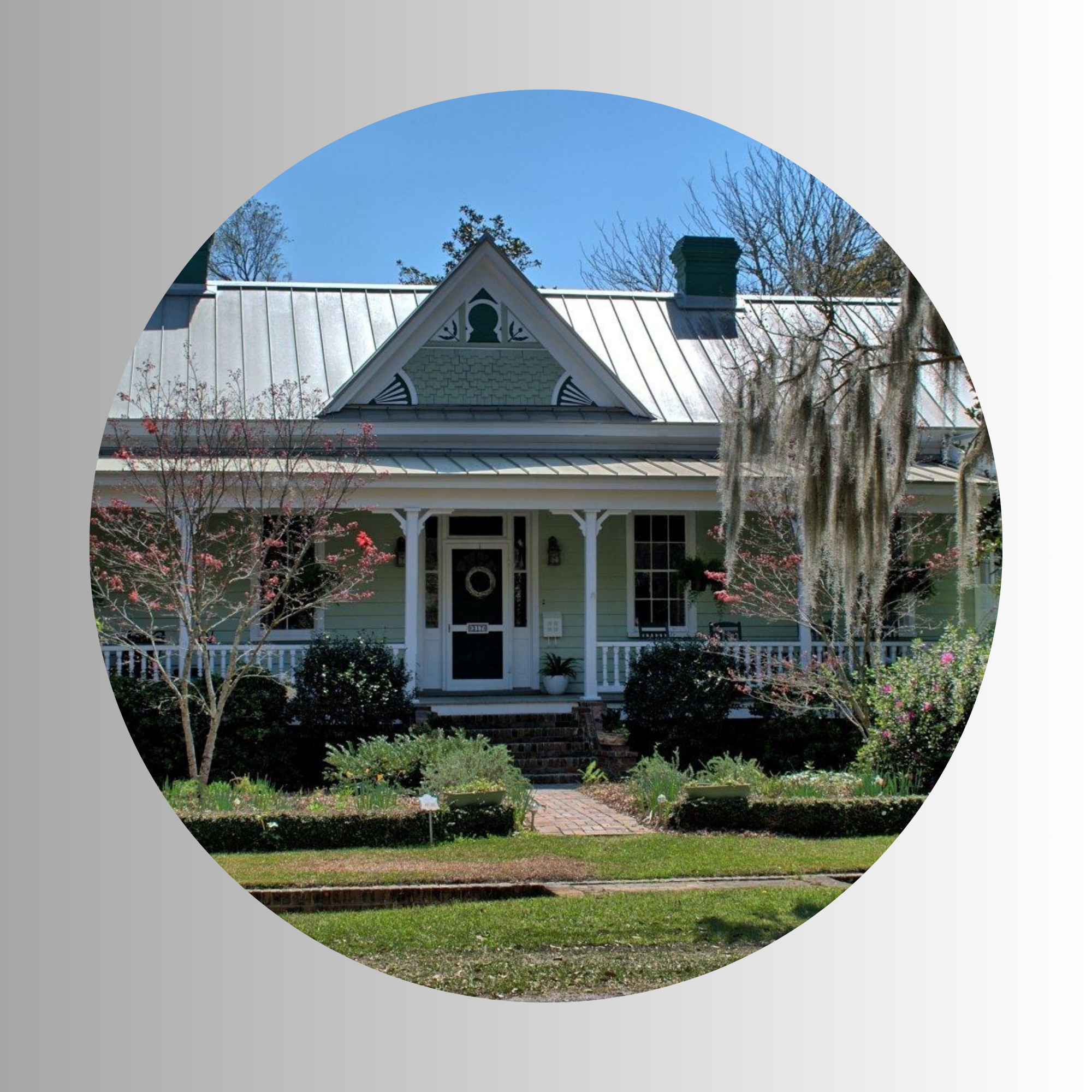 Front view of a house with green walls, white trim, a decorative gable, stairs leading to a porch, surrounded by trees and a lawn.