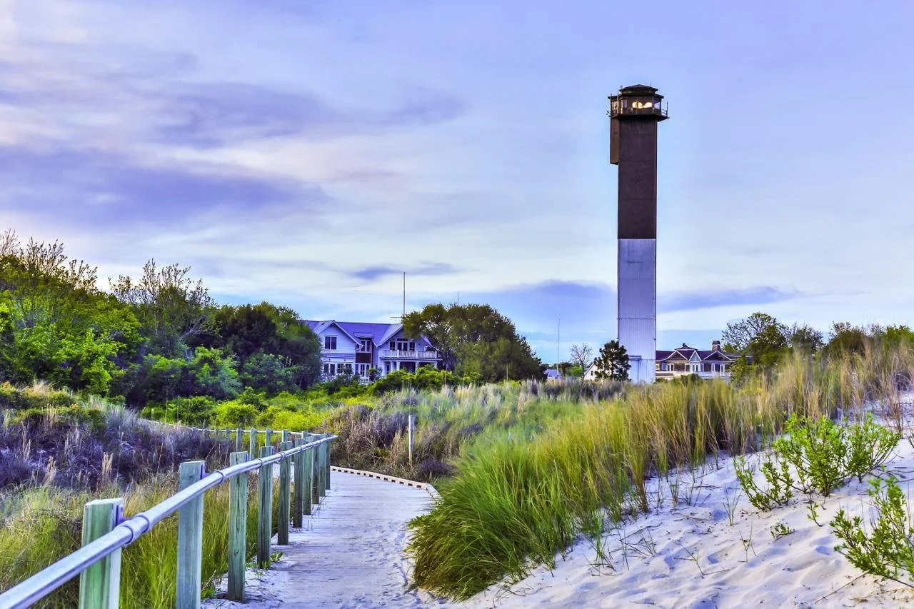A sandy beach pathway with green grass and bushes, leading towards houses and a tall lighthouse in the background under a cloudy sky.