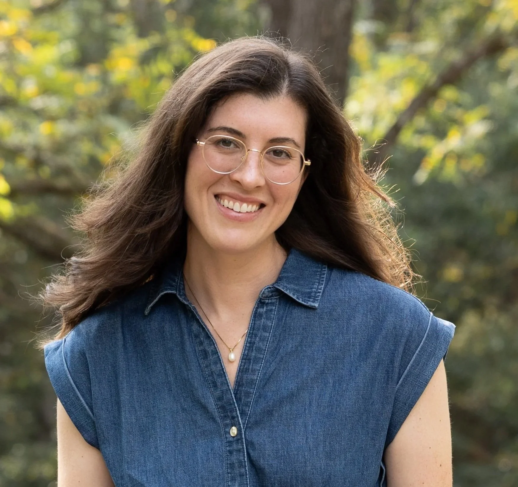 Lori Lockhart, a woman with long brown hair, wearing glasses, a denim shirt, and a pearl necklace, smiling outdoors with blurred green and yellow trees in the background.