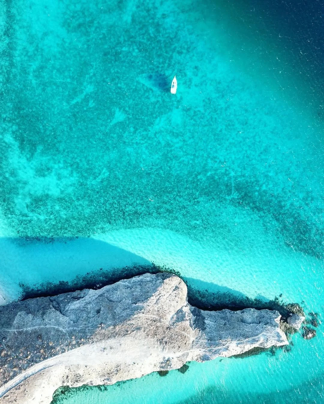 Vista aérea de una playa paradisíaca con agua turquesa transparente, una roca grande en la orilla y una embarcación en el mar.