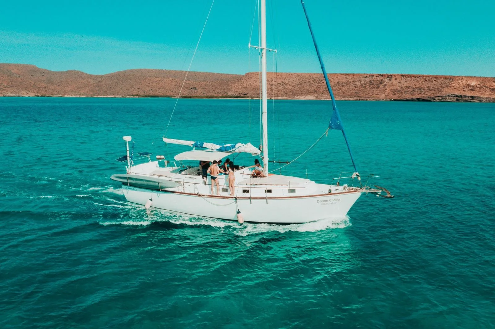 Velero navegando en aguas azules con personas disfrutando, fondo montañas áridas