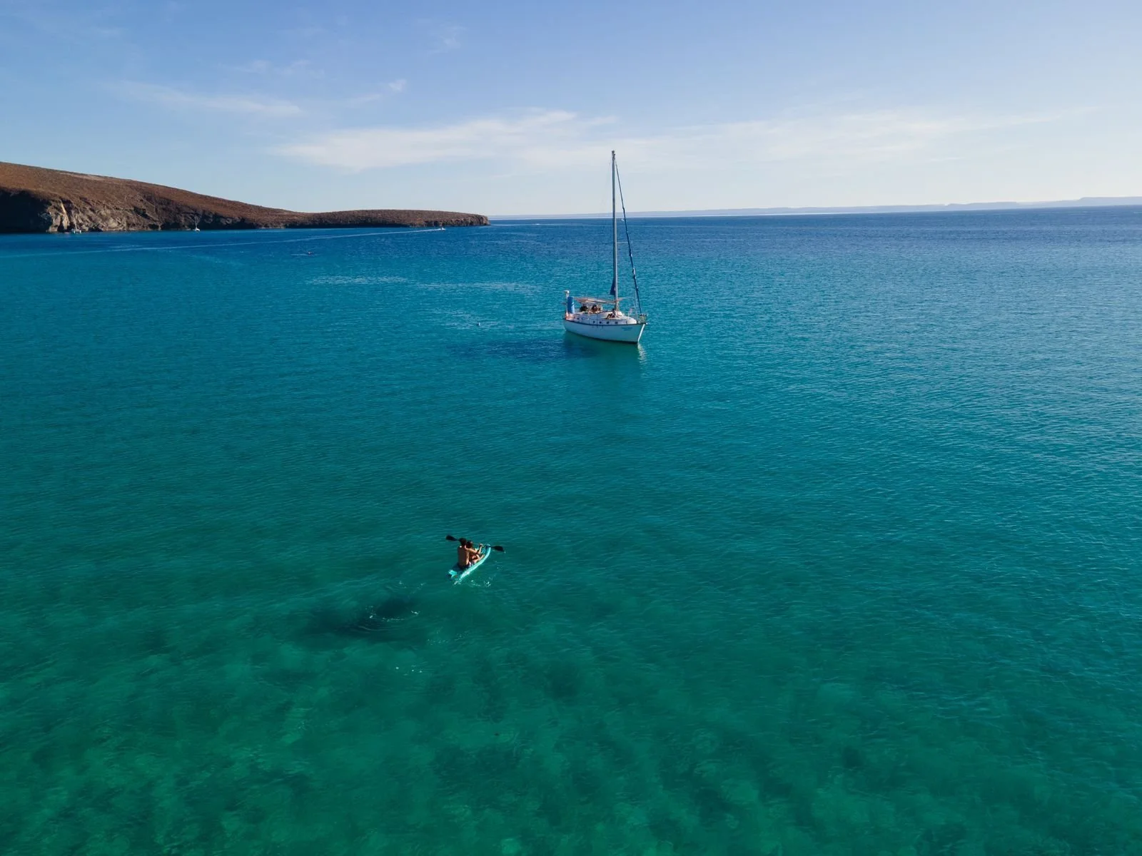 Un velero en el mar, con una persona en kayak en primer plano y un paisaje de costa con colinas en el fondo.