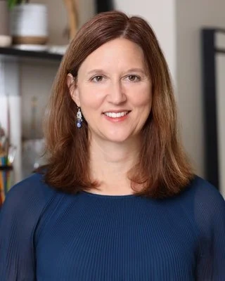 A woman with shoulder-length brown hair, wearing a blue top and earrings, smiling in an indoor setting.