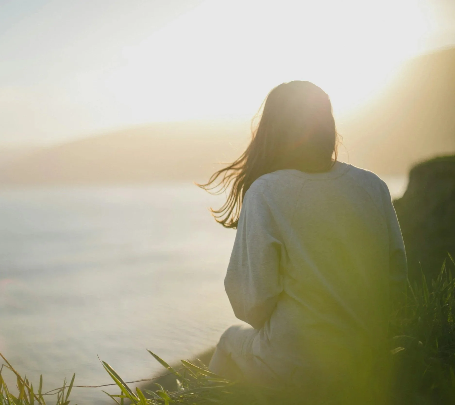 Person with long hair wearing a light-colored hoodie, seen from behind, sitting near water during sunset or sunrise, with reflection and grassy plants in the foreground.