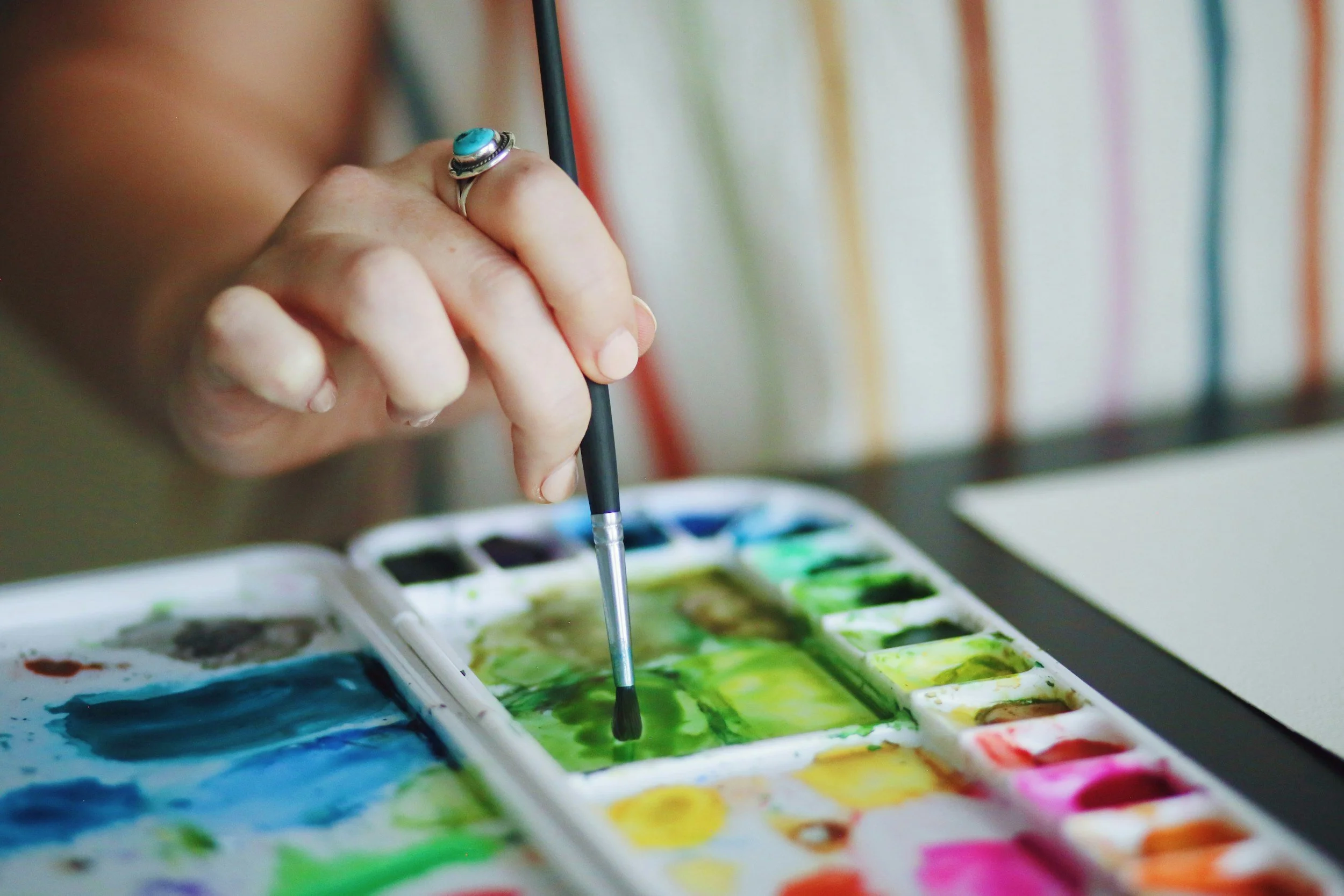 Close-up of a hand holding a paintbrush, dipping into a watercolor palette with various colors.