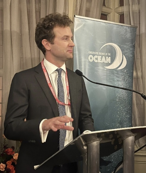 Man in a business suit and tie speaking at a podium with a microphone, during an event promoting ocean conservation.
