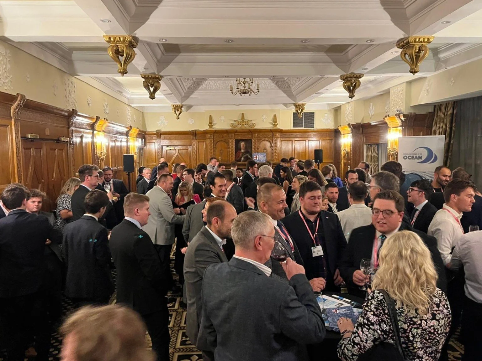 A crowded indoor event with many people in formal attire gathering in a decorated banquet hall, some holding glasses of wine, with wooden paneling and artwork on the walls.