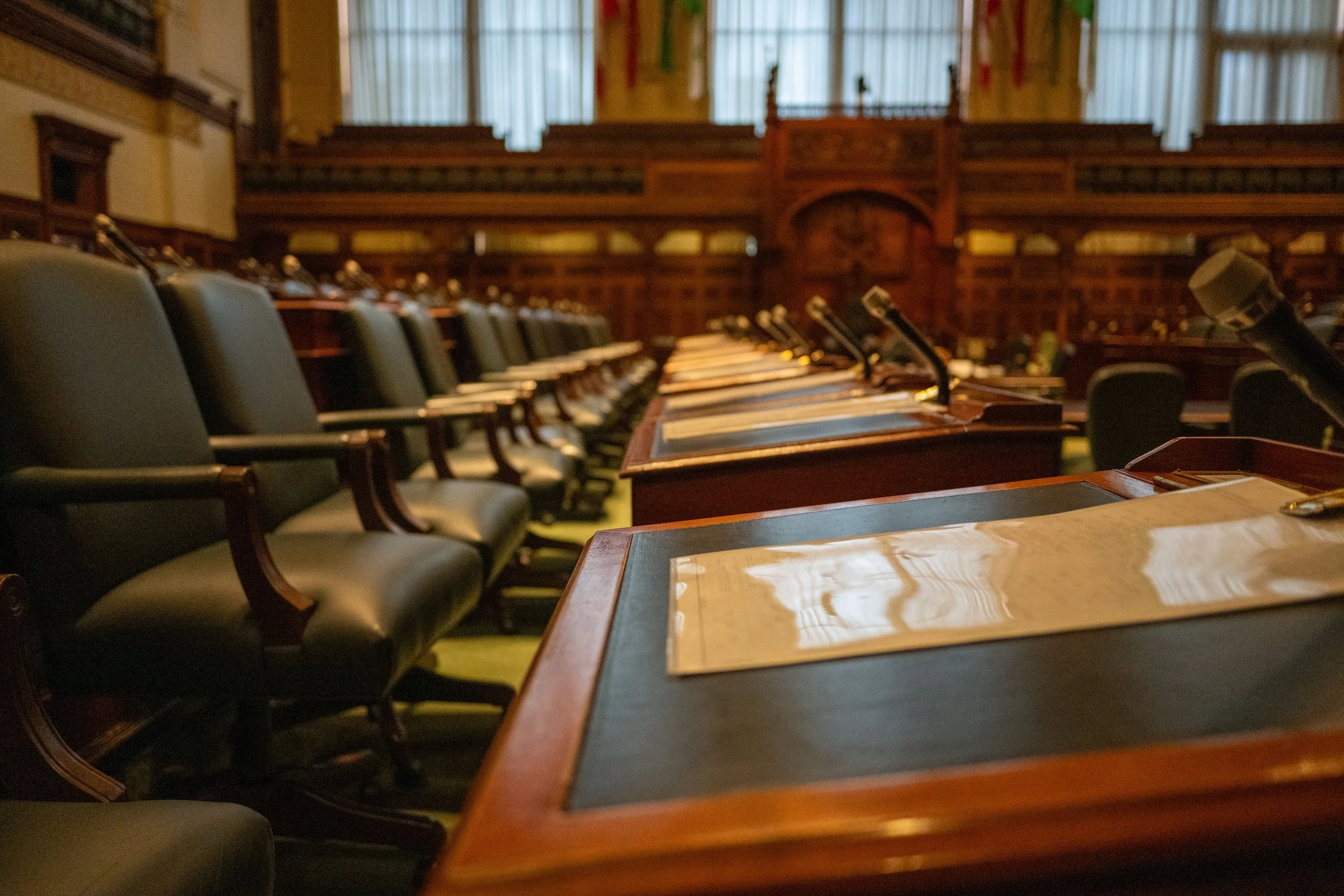 Empty conference room with long wooden tables, black leather chairs, and microphones, in a historic building with wood paneling and high windows.