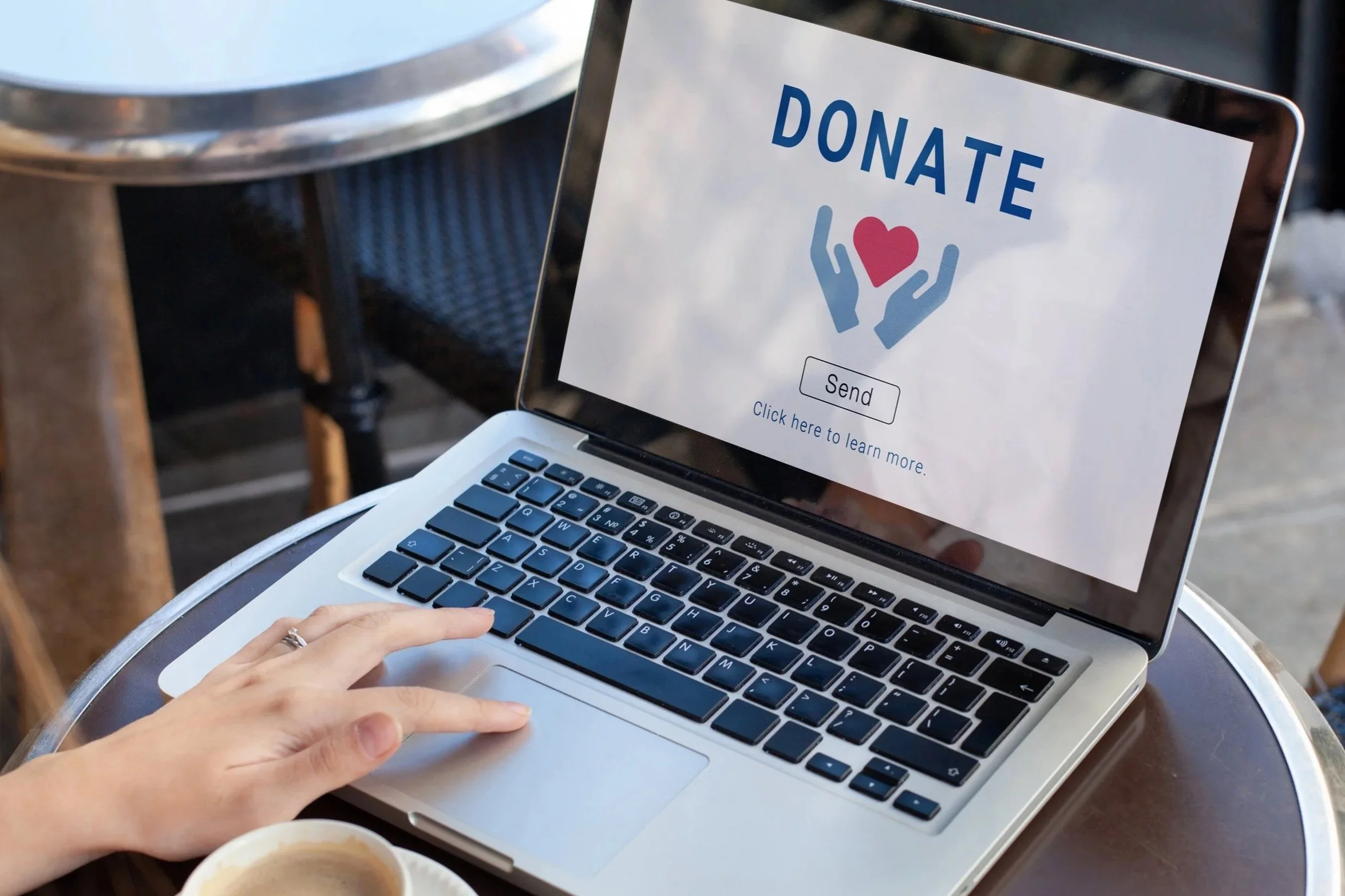 Laptop screen displays a message saying "Donate" with a red heart and blue hands, along with a "Send" button and a link to learn more; a person is typing on the keyboard, and a cup of coffee is partially visible in the foreground.