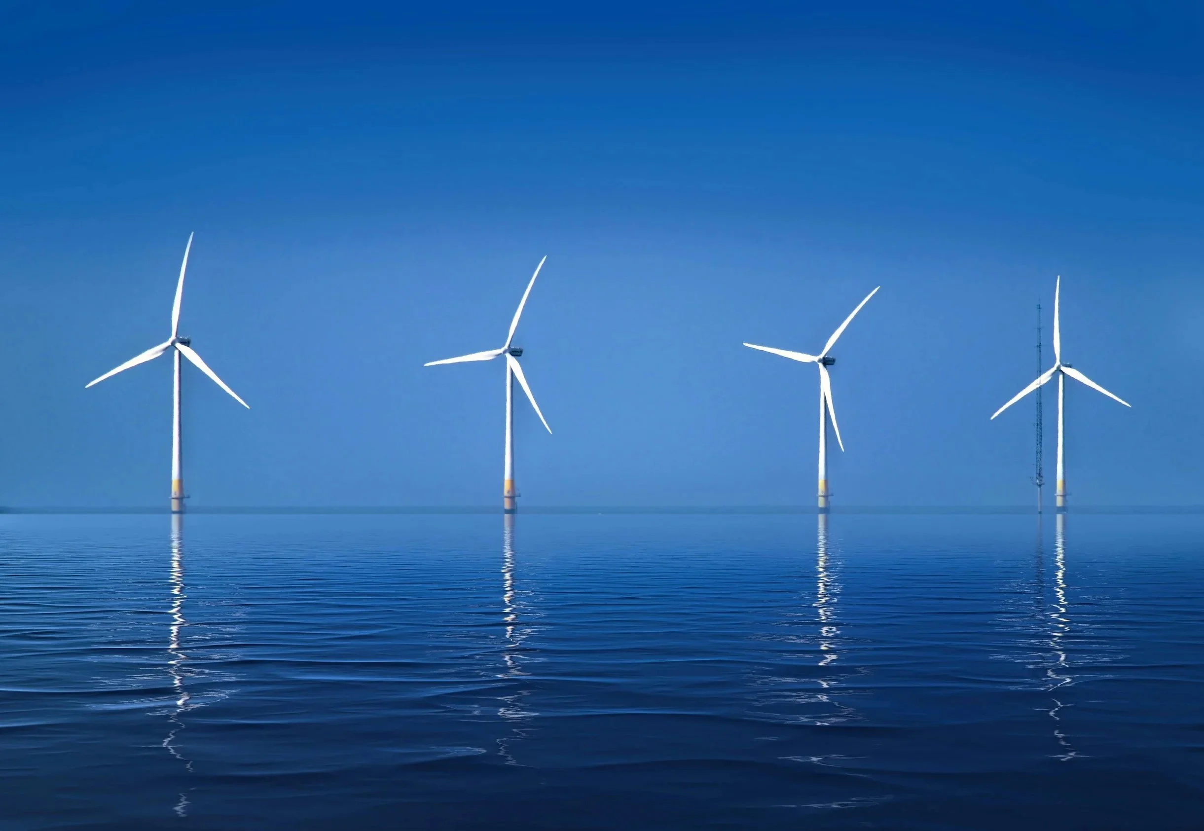Four wind turbines in the ocean, reflected in the water, under a clear blue sky.