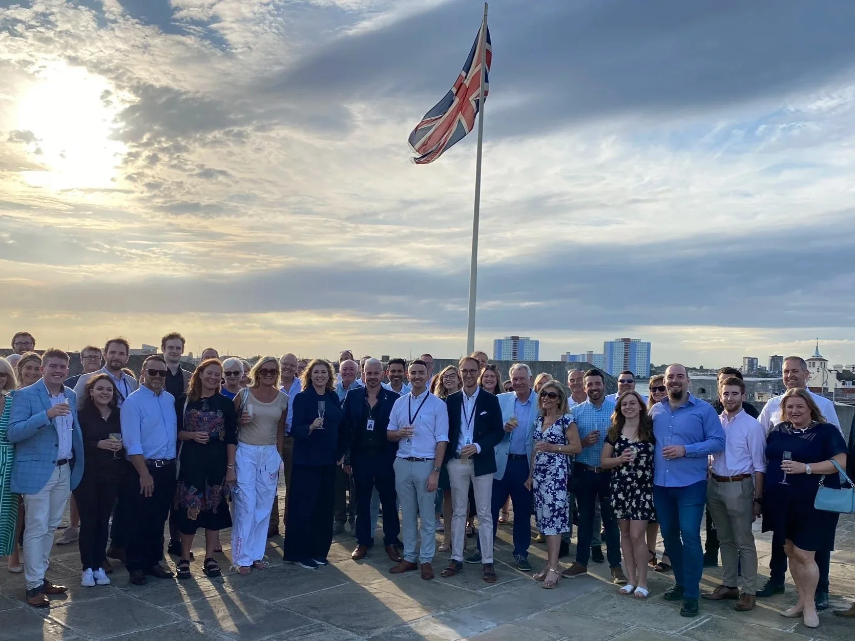 Group of people gathered outdoors on a rooftop during sunset, with a large Union Jack flag flying in the background and a cityscape with tall buildings in the distance.
