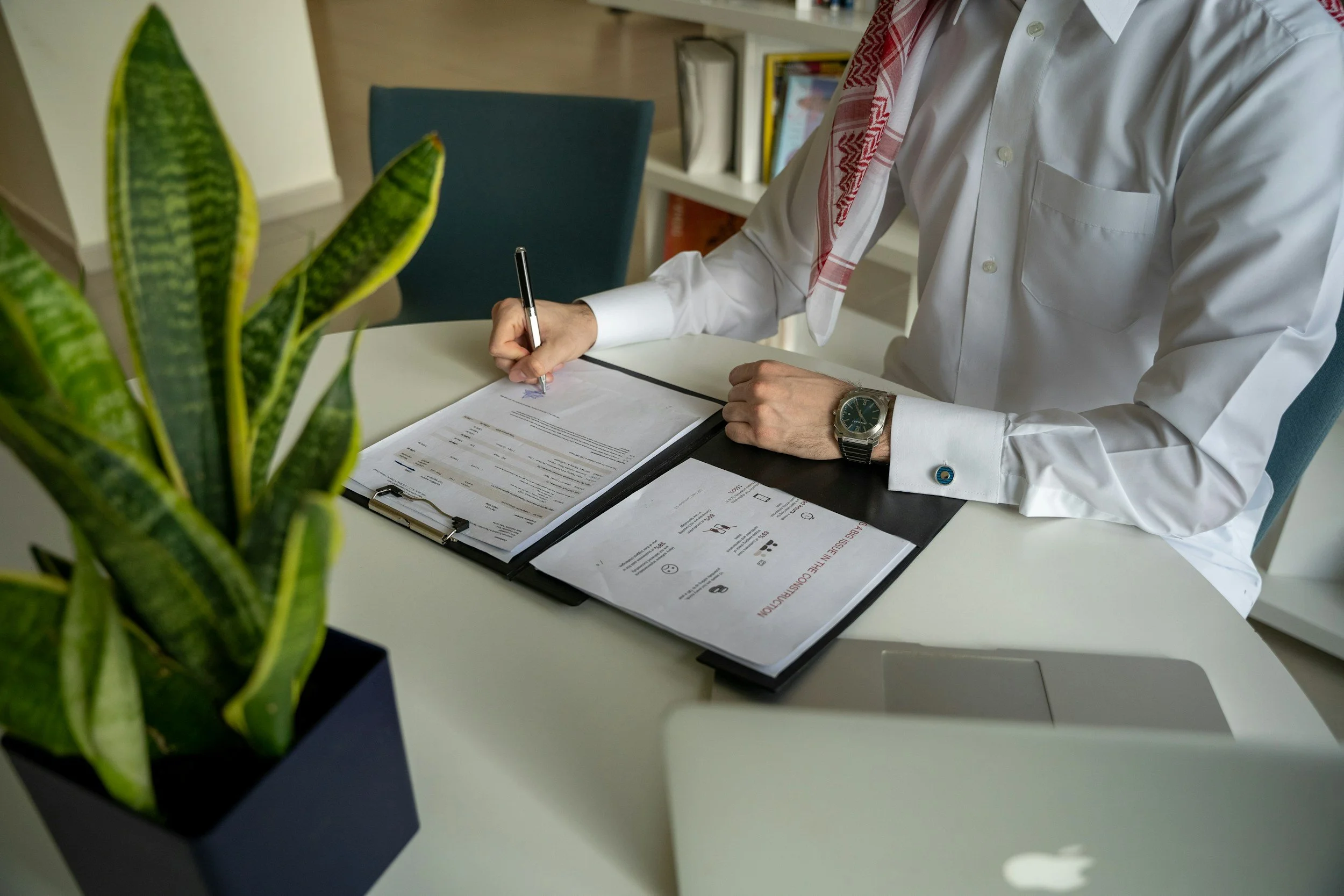 Personne en chemise blanche, portant un foulard rouge et blanc, assise à un bureau en train de signer un document, avec des livres sur une étagère en arrière-plan, et une plante verte en premier plan à gauche.