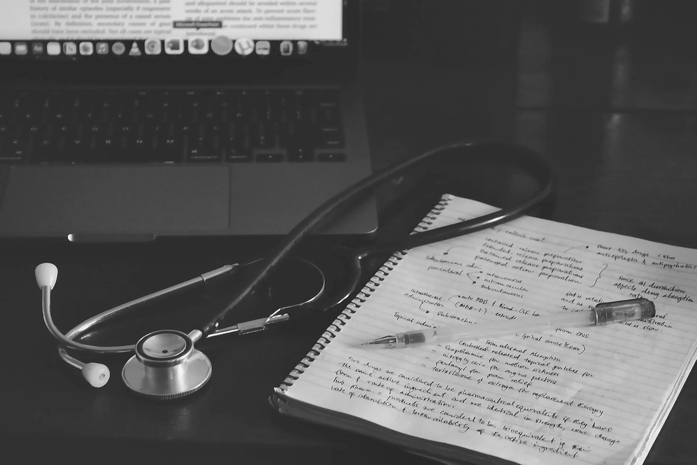 Une table de travail avec un ordinateur portable allumé, un stéthoscope, des écouteurs, un cahier avec des notes manuscrites et un stylo.