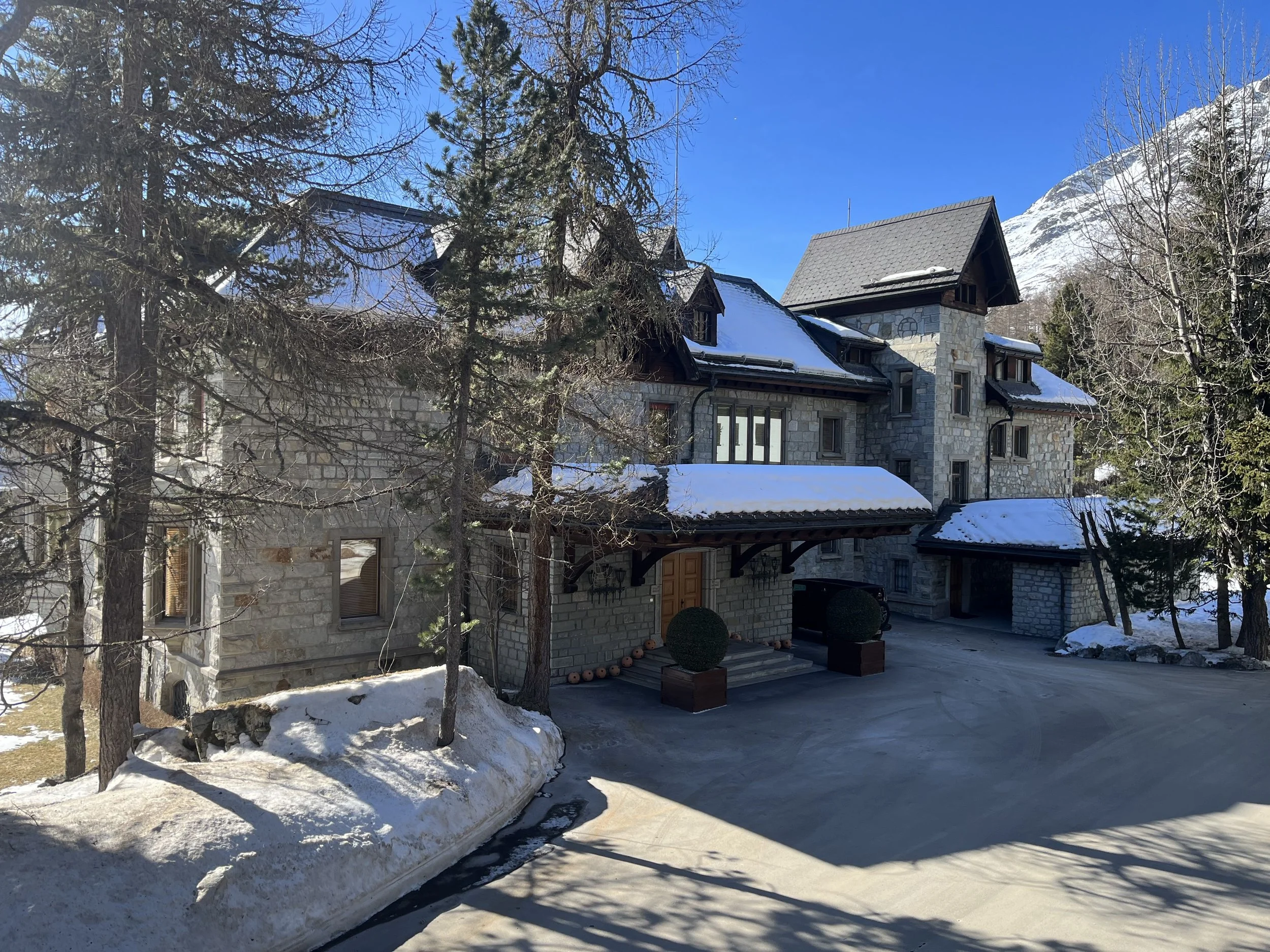 A stone house with multiple peaked roofs in a snowy, mountainous landscape, surrounded by leafless trees and evergreens with snow on their branches, under a bright blue sky.