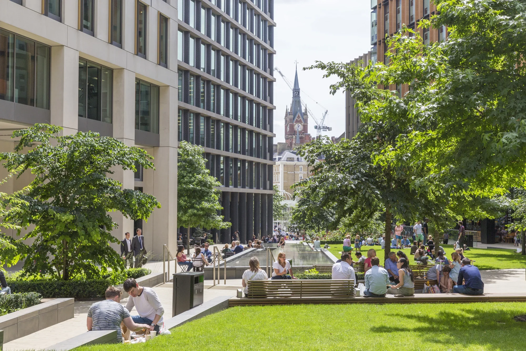 A busy urban park with many people sitting, walking, and relaxing on benches and grass. Surrounding greenery with trees and shrubs, modern buildings, and a historic church with a tall steeple in the background.
