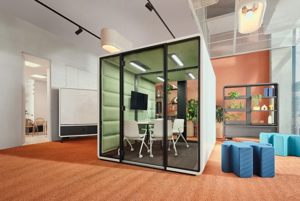 Modern office space with a glass-enclosed meeting pod, white chairs, a wall-mounted TV, and indoor plants, featuring orange carpeting and contemporary decor.