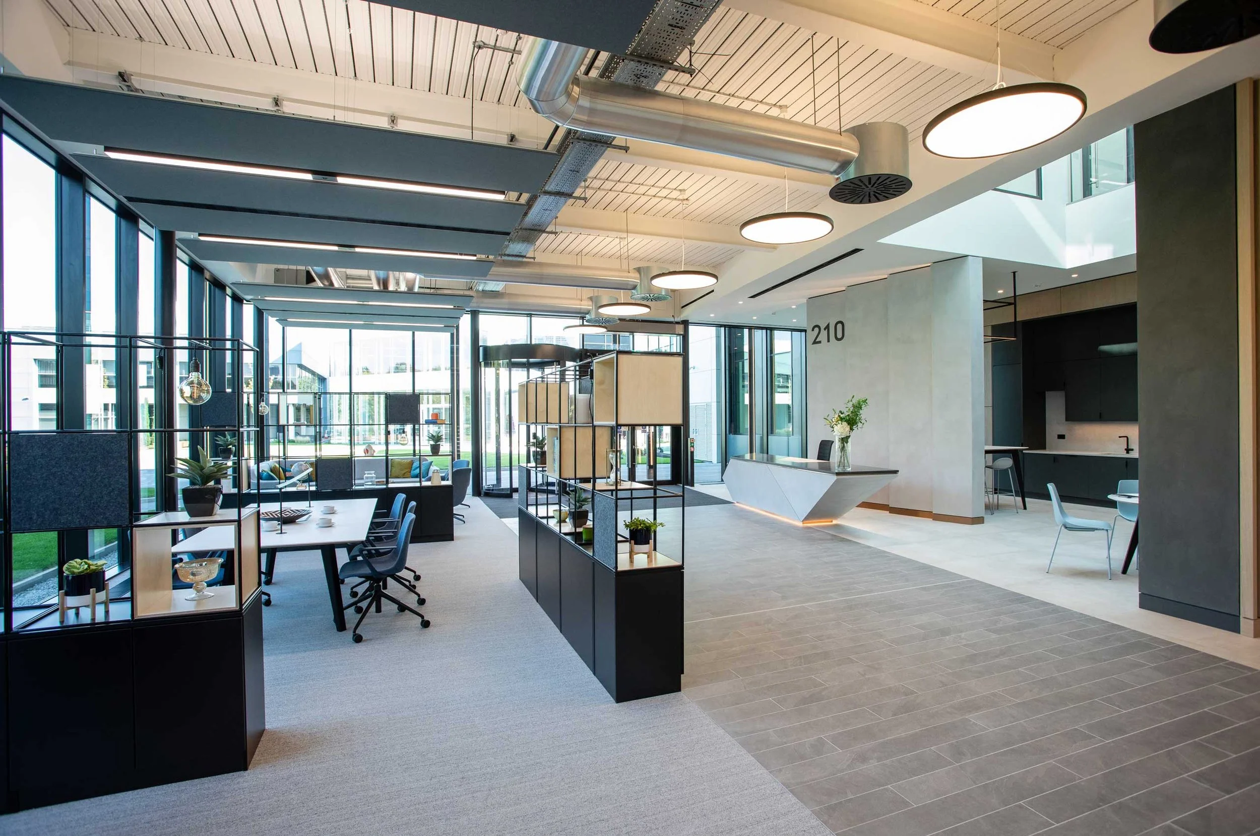 Modern office lobby with large windows, high ceiling with exposed ductwork, round ceiling lights, a reception desk, seating area, and decorative shelves with plants.