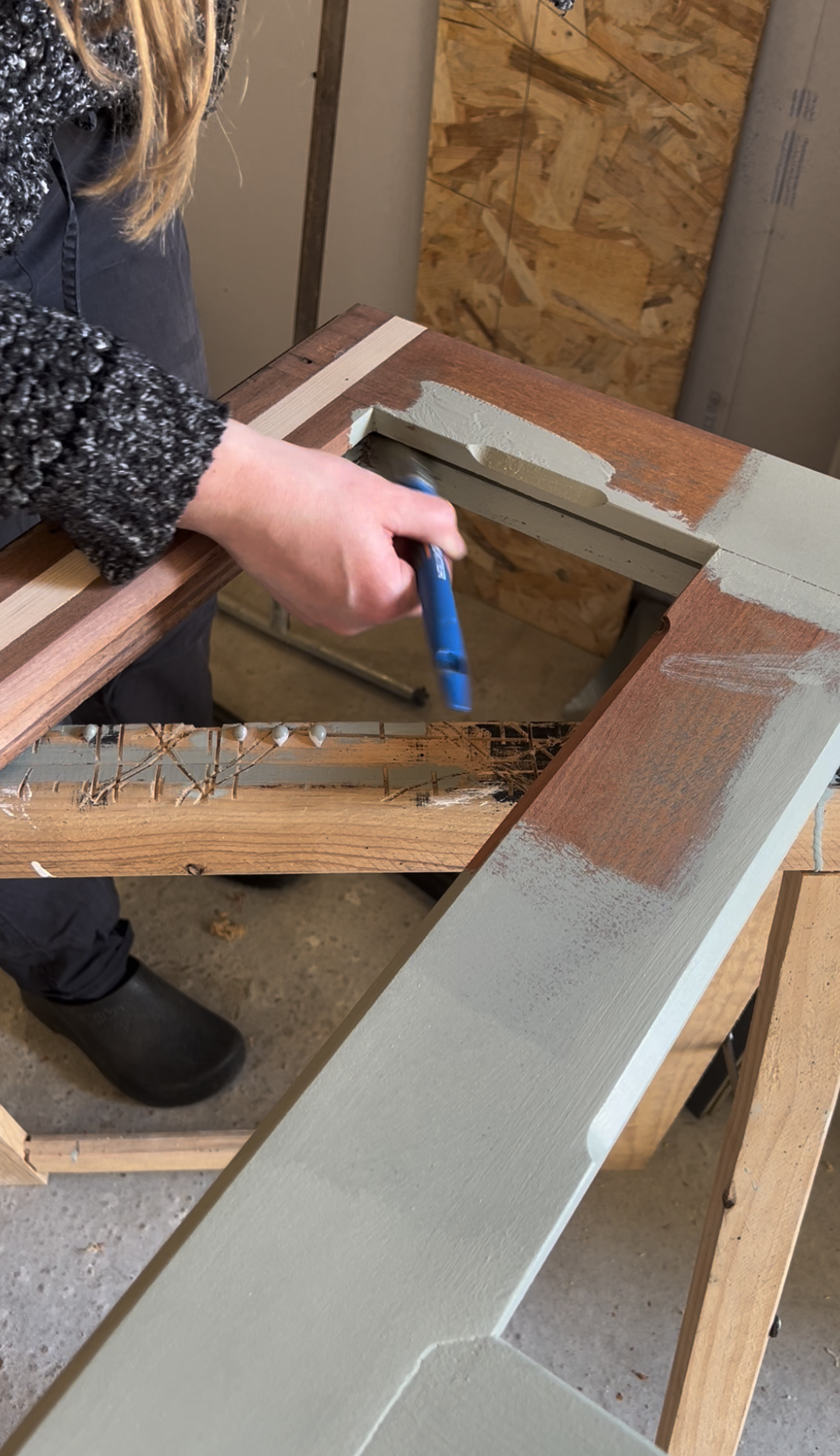 Une personne peint une structure en bois avec de la peinture beige. La personne porte des bottes noires et un pull gris. L'image montre un espace de travail avec des murs en contreplaqué.
