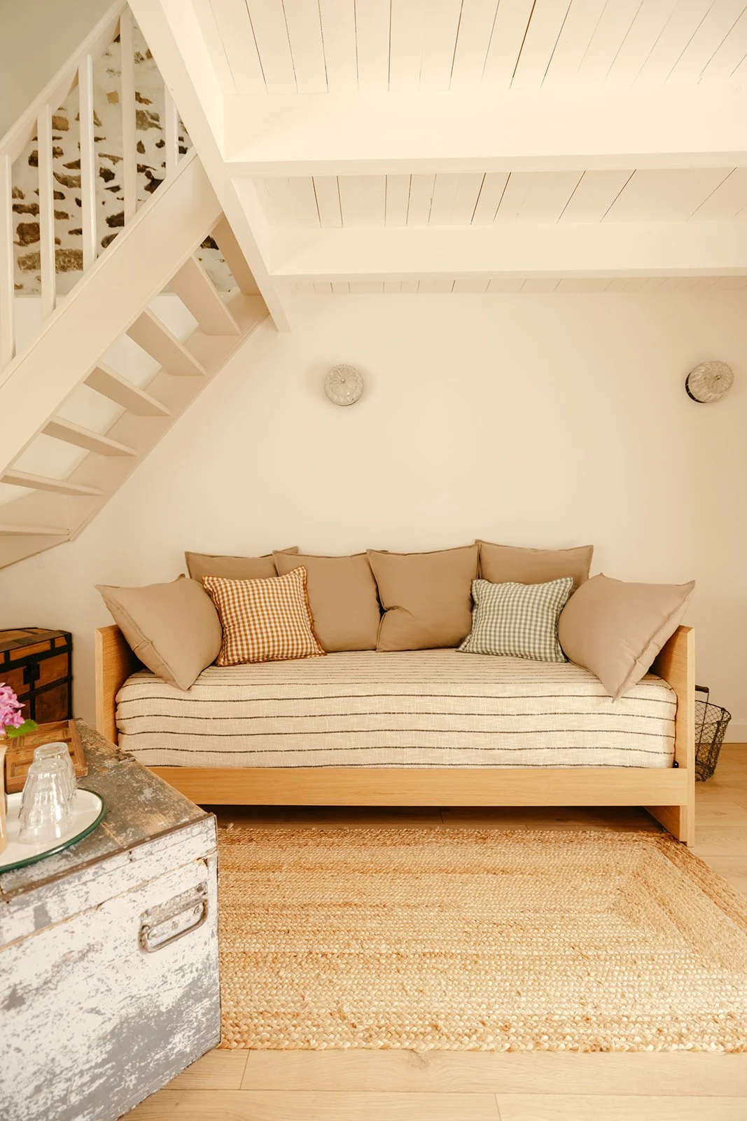Un canapé beige avec des coussins à carreaux et un tapis en jute dans un salon avec un plafond en bois peint en blanc et deux lampes suspendues.