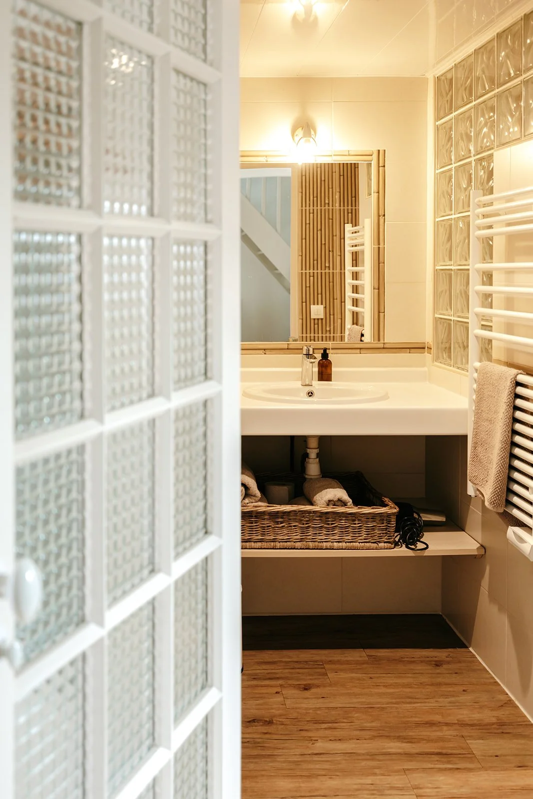 Vue partielle d'une salle d'eau avec lavabo, miroir, radiateur électrique, et décoration en bois, vue à travers une porte en verre dépoli.