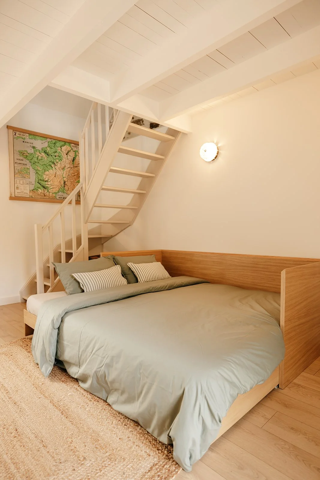 Chambre Padine du Clos des Sentiles à Perros-Guirec avec un lit double en bois, une couette vert pâle, des oreillers rayés, un mur blanc, un escalier menant à la mezzanine, une ancienne carte scolaire vintage accrochée au mur et des appliques.