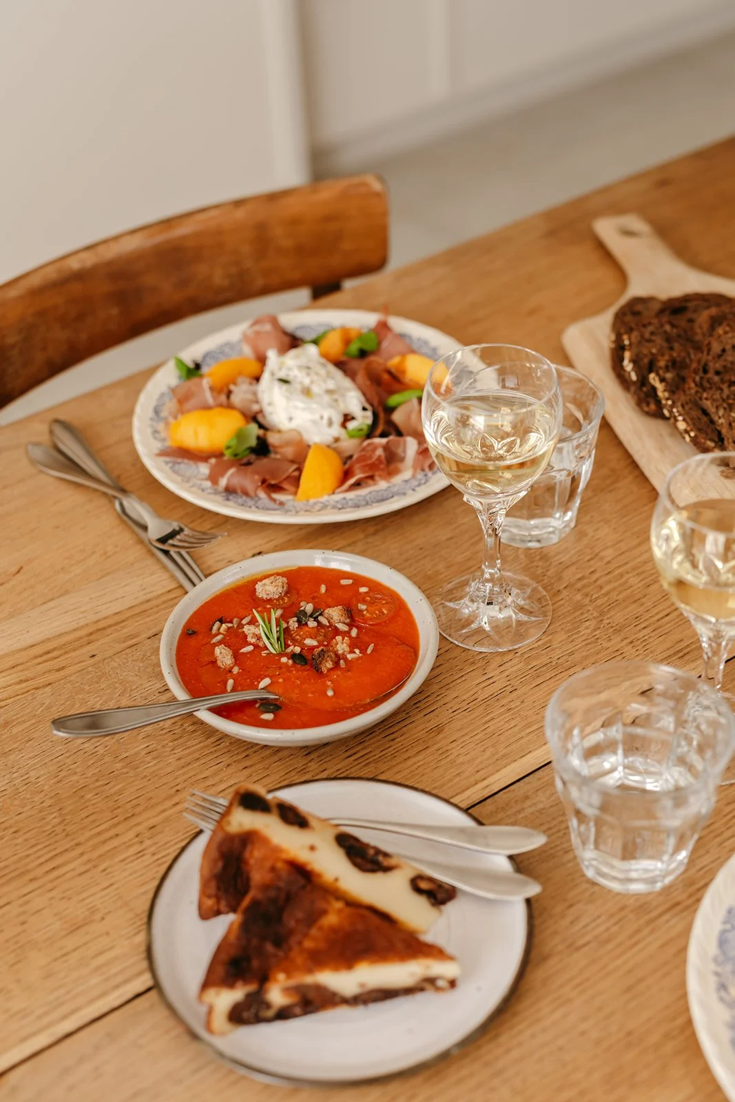 Une table en bois avec une salade, une soupe à la tomate, des verres de vin blanc, et un morceau de far breton.