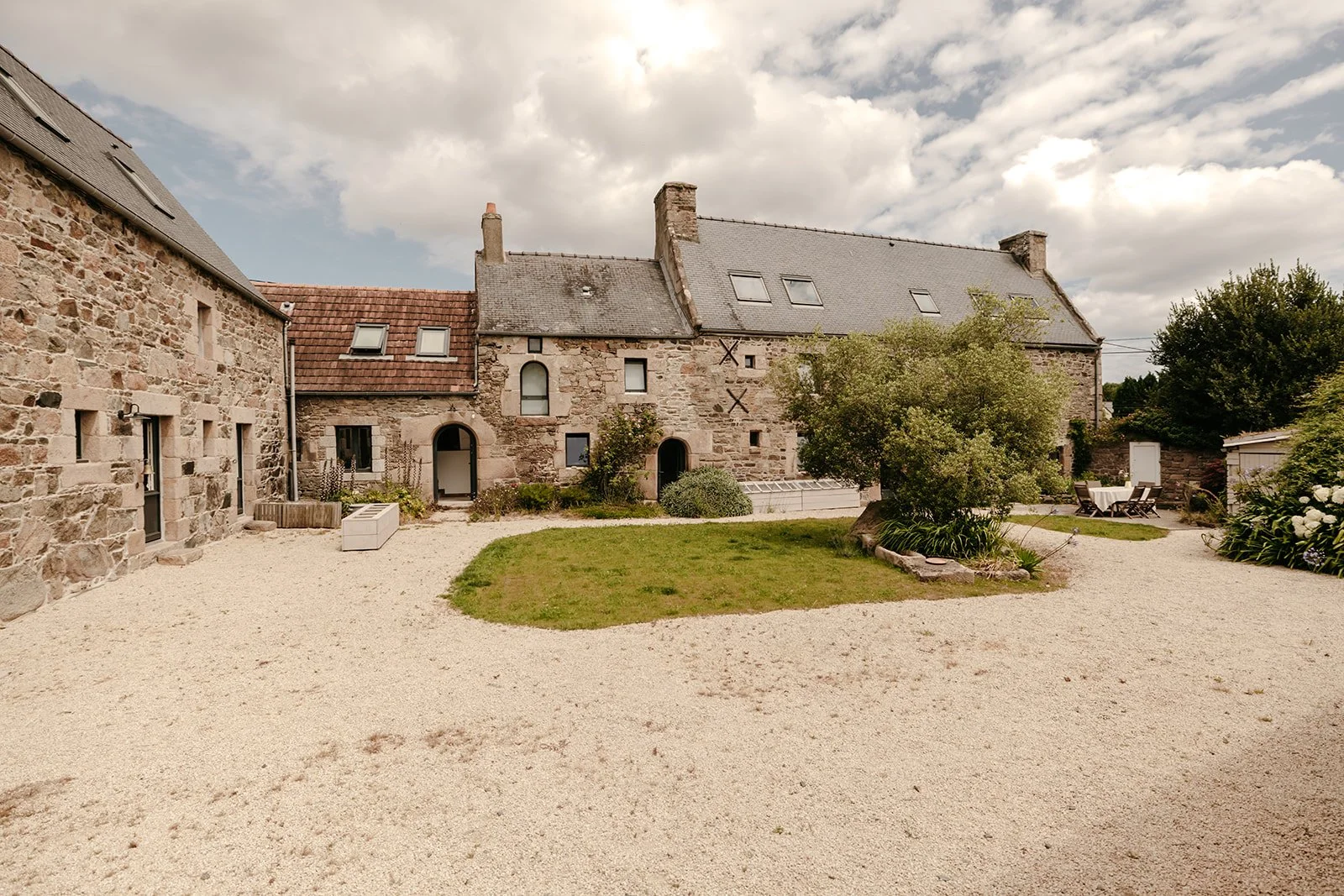 Cour extérieure d'une ferme en pierre rénovée avec un ancien olivier au milieu de la cour et un chemin en gravier.
