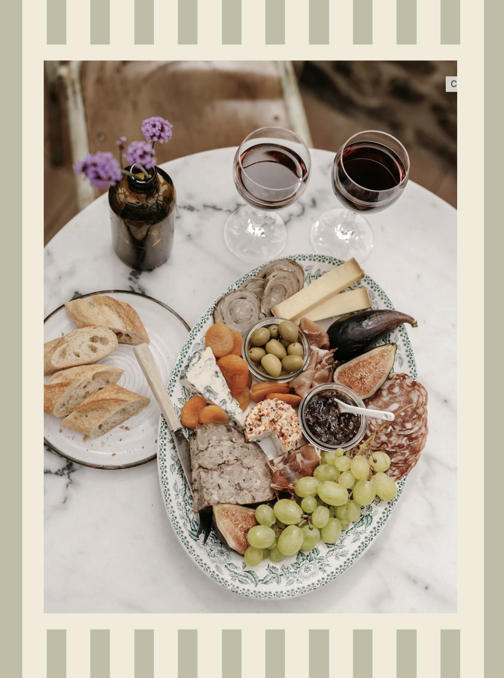 Plate de charcuterie avec fromage, fruits, olives, pain, et dips, accompagnée de deux verres de vin rouge sur une table en marbre.