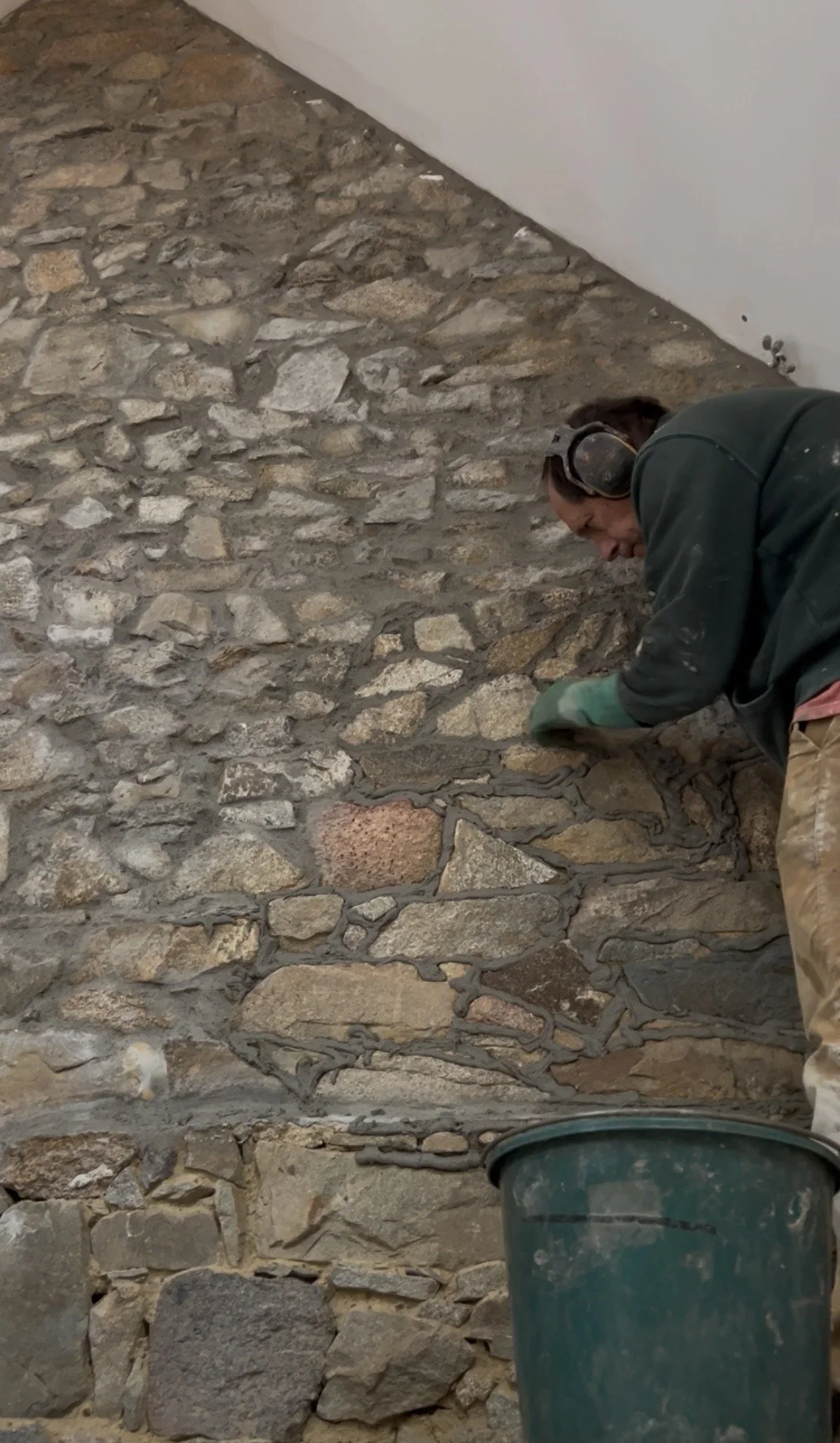 Un homme travaille sur un mur en pierre avec un masque antibruit dans une pièce en cours de rénovation.