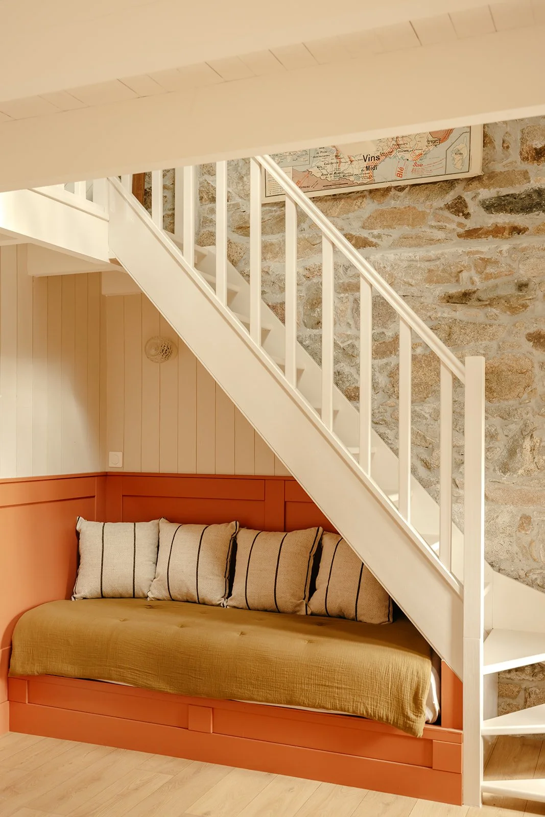 Banquette intégrée sous un escalier en bois peint blanc avec coussins, mur en pierre et carte scolaire vintage accrochée au mur.